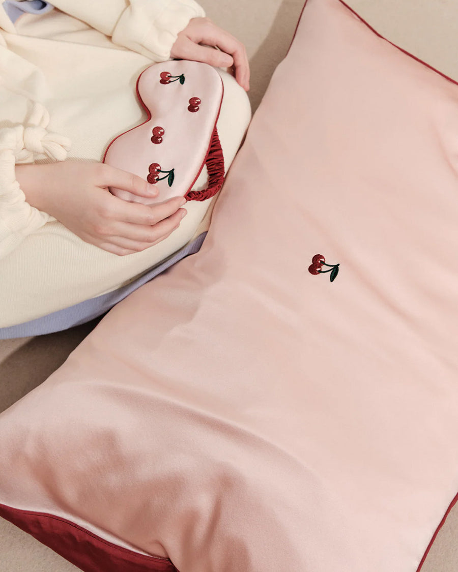 model sitting with light pink mulberry silk eye mask with embroidered all over cherry print and pink pillowcase with dainty embroidered cherries on the center
