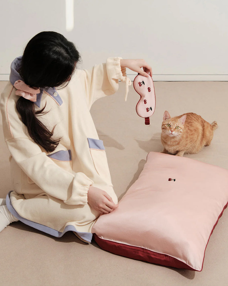 model sitting with light pink mulberry silk eye mask with embroidered all over cherry print and pink pillowcase with dainty embroidered cherries on the center