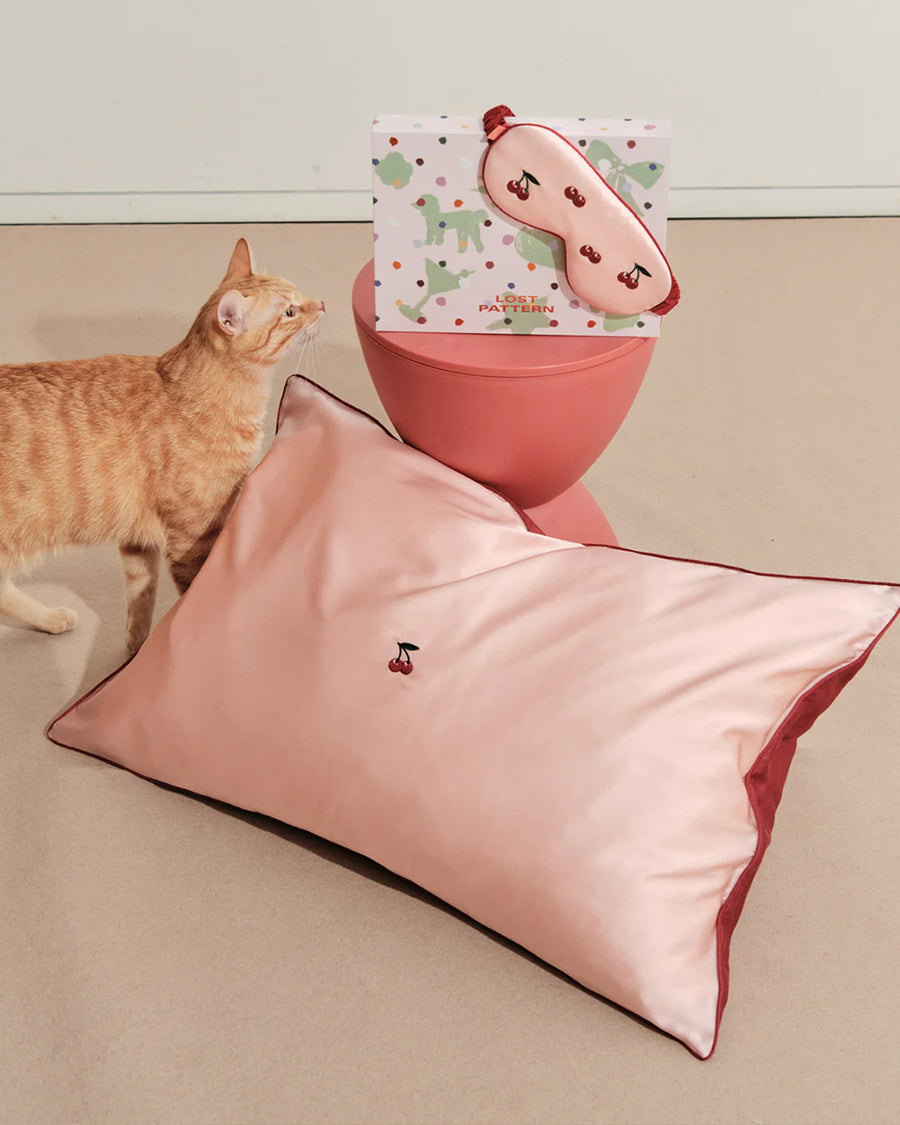 light pink mulberry silk eye mask with embroidered all over cherry print and pink pillowcase with dainty embroidered cherries on the center with an orange cat next to it