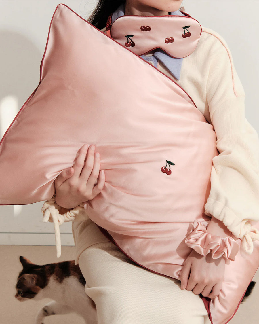 model holding light pink mulberry silk eye mask with embroidered all over cherry print and pink pillowcase with dainty embroidered cherries on the center and pink scrunchie