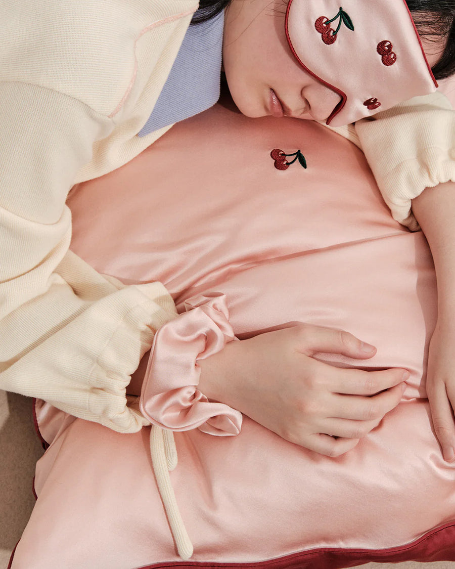 model using light pink mulberry silk eye mask with embroidered all over cherry print and pink pillowcase with dainty embroidered cherries on the center and pink scrunchie