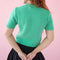 back view of model wearing pale green short sleeve mockneck sweater with white cursive 'stay soft' across the front