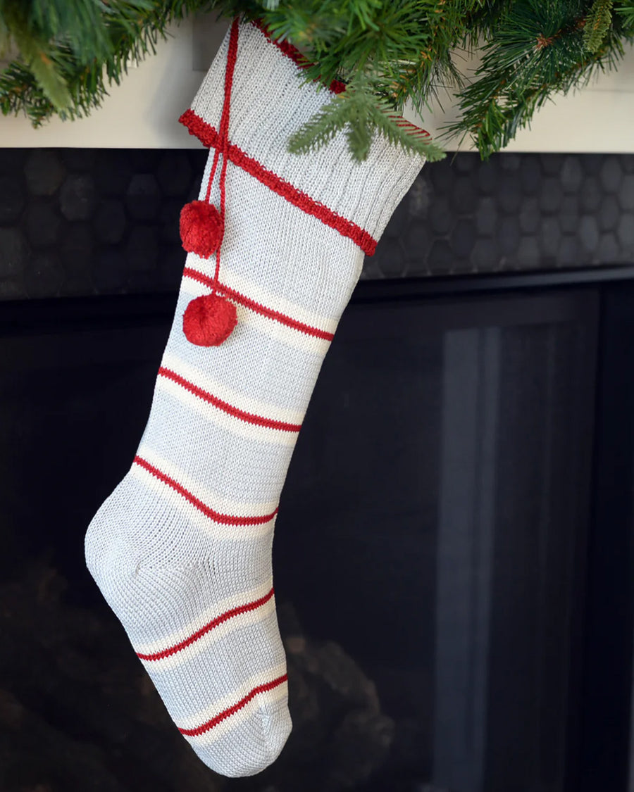 grey cotton stocking with red and white thin stripes, red trim on the cuff and red pom pom detail hanging on a fireplace
