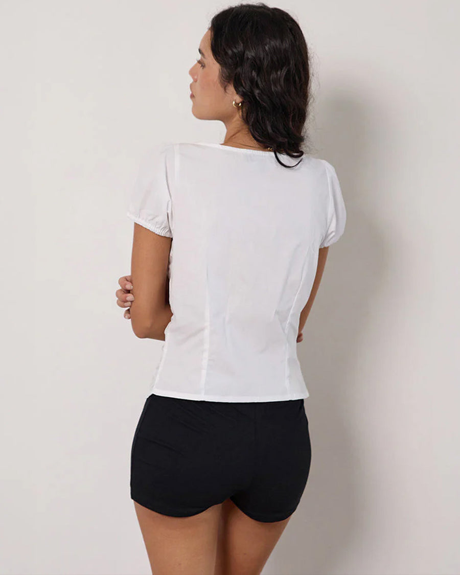 back view of model wearing white cotton short sleeve blouse with deep v-neck, button front and corset waist and black shorts