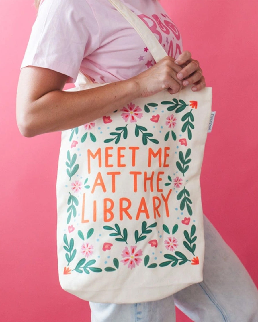 model holding tan canvas bag with orange 'meet me at the library' surrounded by colorful flowers