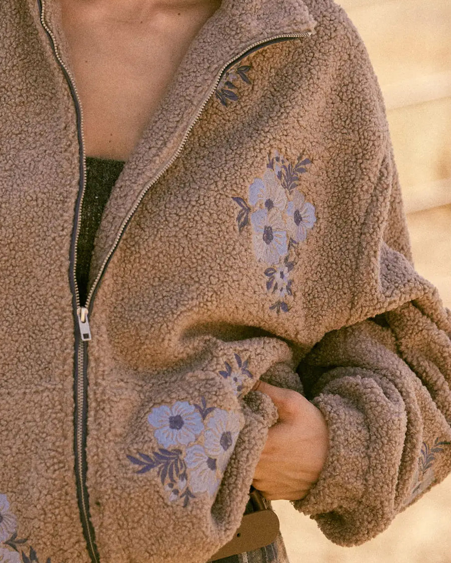 up close of model wearing tan sherpa zip-up jacket with purple embroidered clustered flowers
