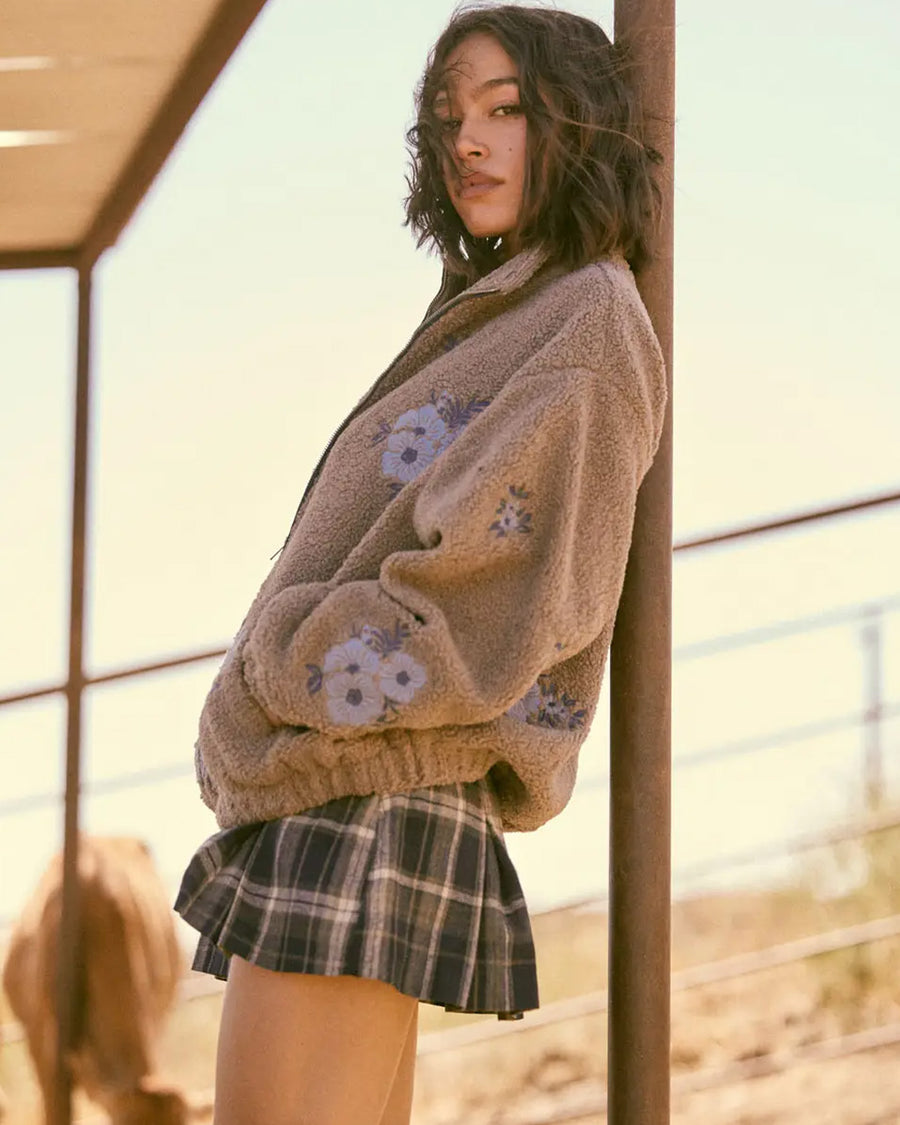 side view of model wearing tan sherpa zip-up jacket with purple embroidered clustered flowers