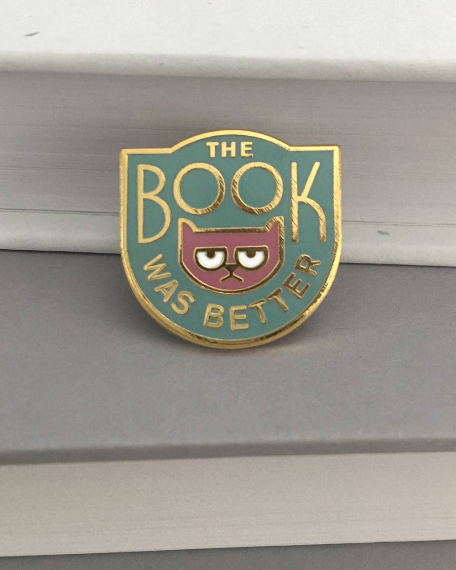 teal enamel pin with gold 'the book was better' across the front and cat graphic
