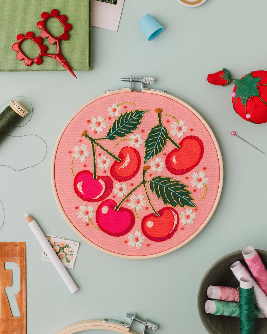 pink embroidery kit with all over cherry and white flowers on a blue ground surrounded by crafting items