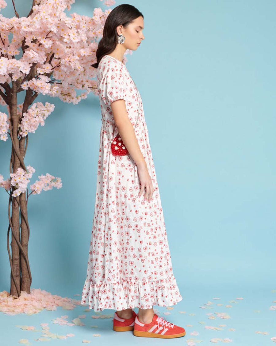 side view of model wearing white maxi dress with red heart and flower embroidery, ruffle hem and puff short sleeves