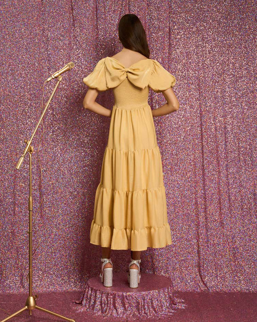 back view of model wearing golden yellow tiered midi dress with puff sleeves, smocked bodice, and back oversized bow