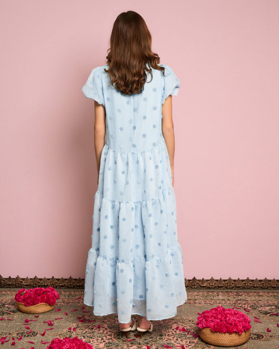 backview of model wearing light blue tiered midi dress with puff sleeves and subtle pom detail
