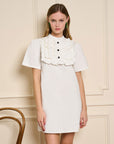 model wearing white mini dress with ruffle bib front with black buttons and short sleeves