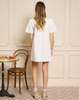 back view of model wearing white mini dress with ruffle bib front with black buttons and short sleeves