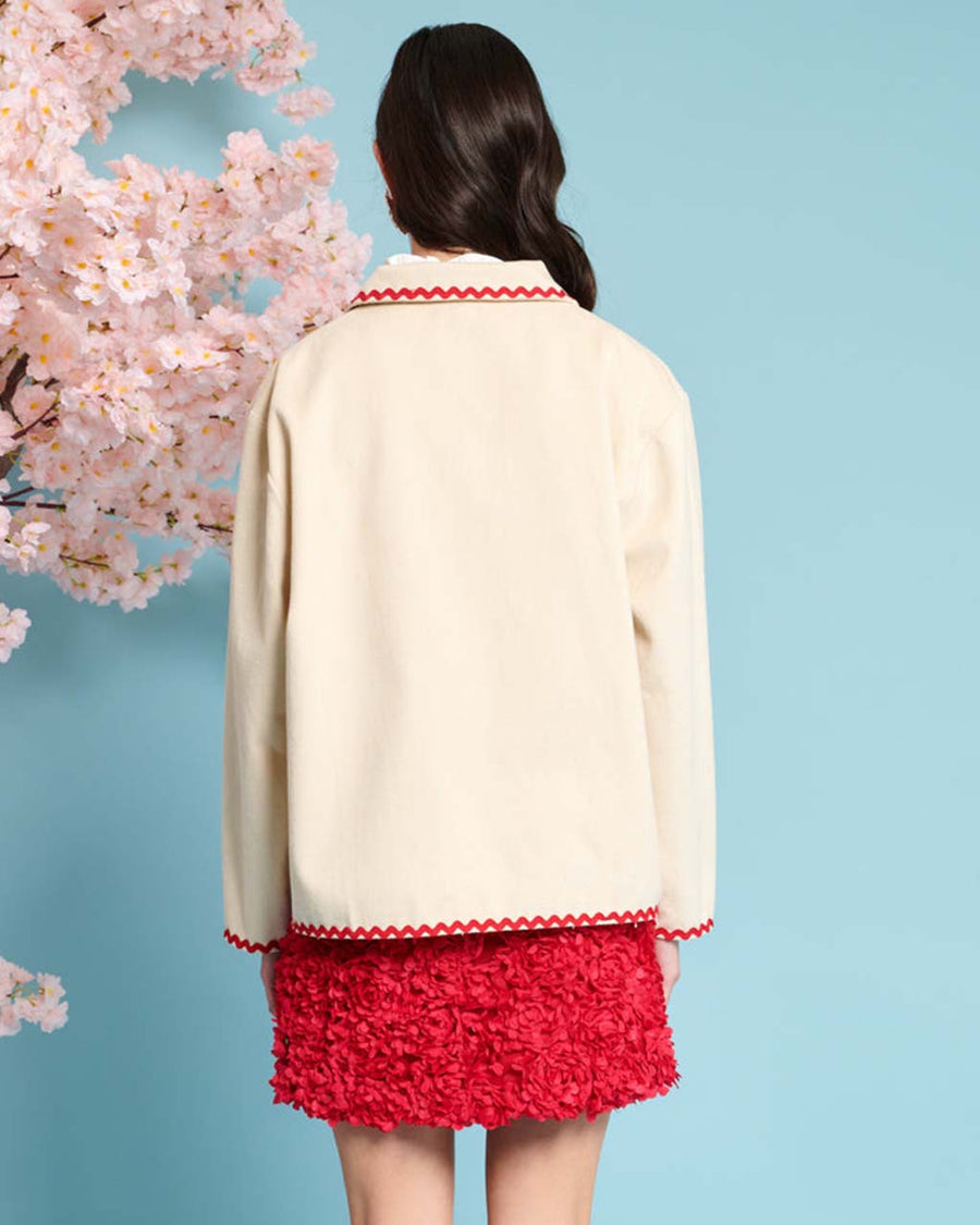 back view of model wearing natural jacket with red ric rac trim and embroidered heart detail on front patch pockets