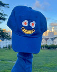 royal blue baseball cap with smiley with egg eyes and banana mouth