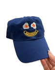model holding royal blue baseball cap with smiley with egg eyes and banana mouth
