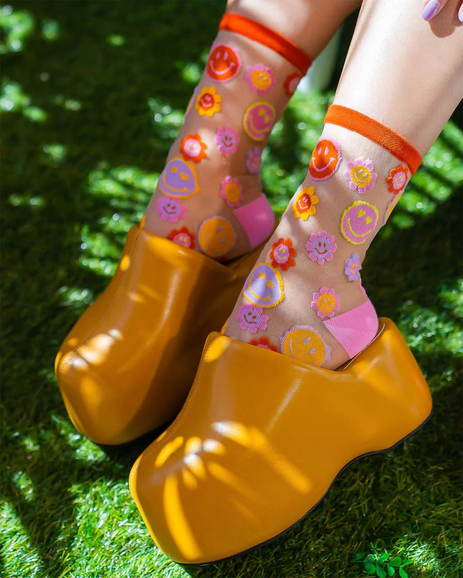 model wearing sheer socks with colorful smiley face and smiley floral print and yellow chunky clogs