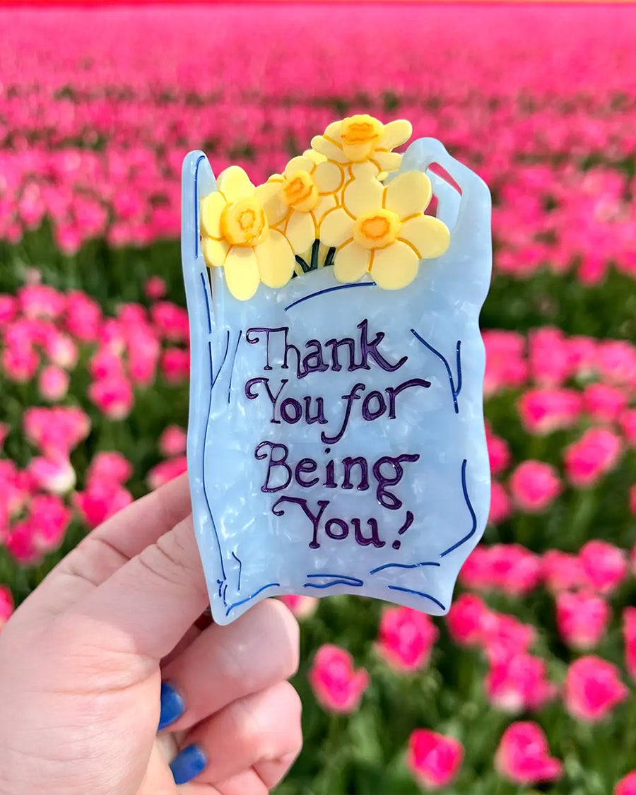 model holding light blue 'plastic bag' hair claw with 'thank you for being you' across the front and yellow daffodils coming from the top