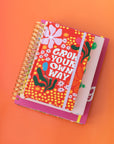 17-month classic planner with red ground, colorful abstract flowers and white 'grow your own way' text across the front on a stack of notebooks