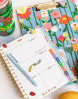 clipboard folio with blue ground and colorful abstract floral print with planner, pen and tumbler next to it