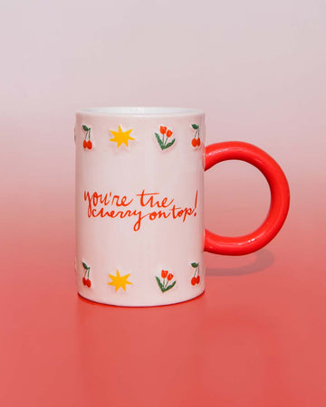 White mug with red handle, with cherry, start and tulip illustrations, that says "you're the cherry on top!" on a red gradient background