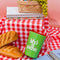 green small steel tumbler with 'life's a picnic' and pink lid on red and white checkered blanket surrounded by bread, planners, and a basket with fruit and pens inside