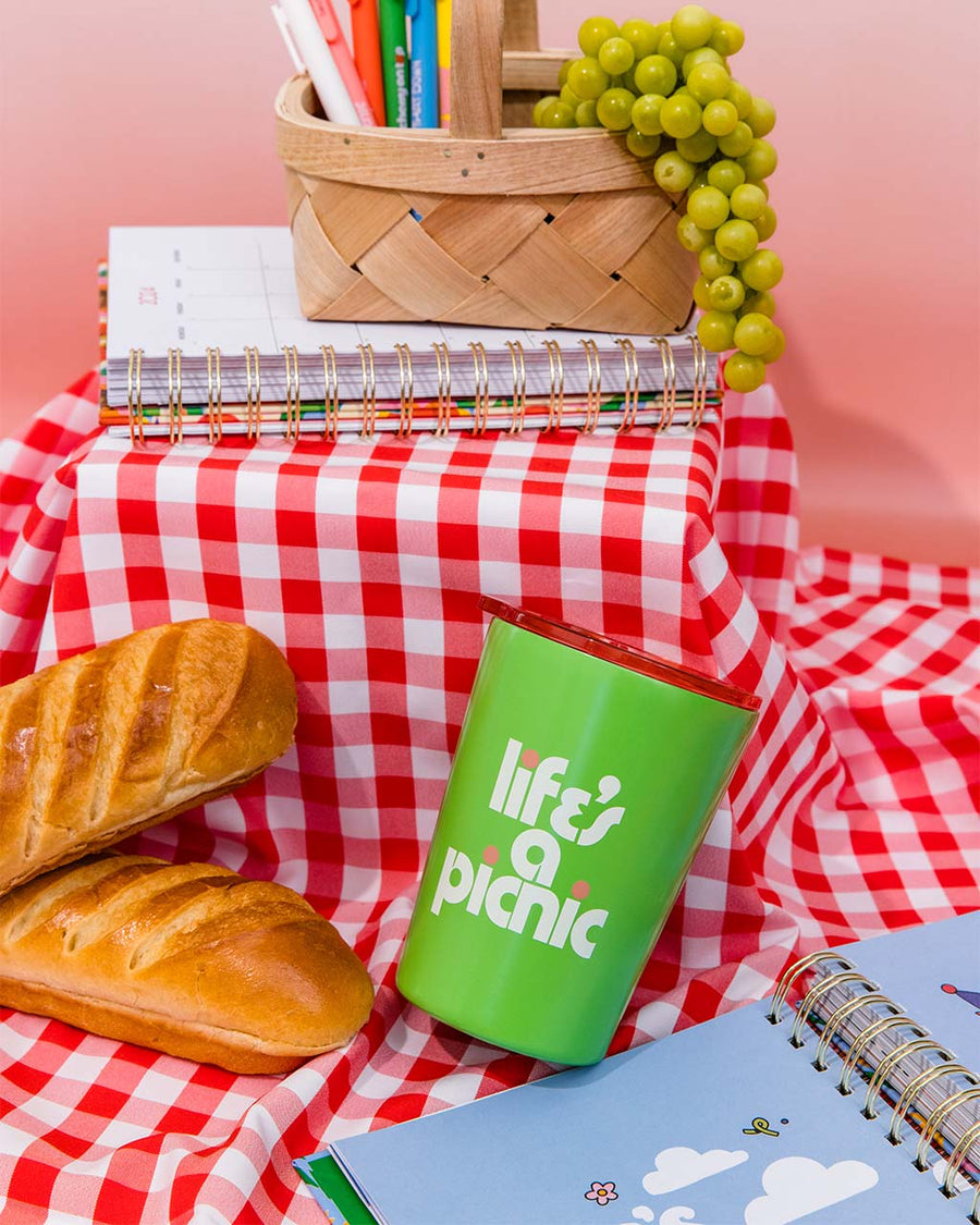green small steel tumbler with 'life's a picnic' and pink lid on red and white checkered blanket surrounded by bread, planners, and a basket with fruit and pens inside