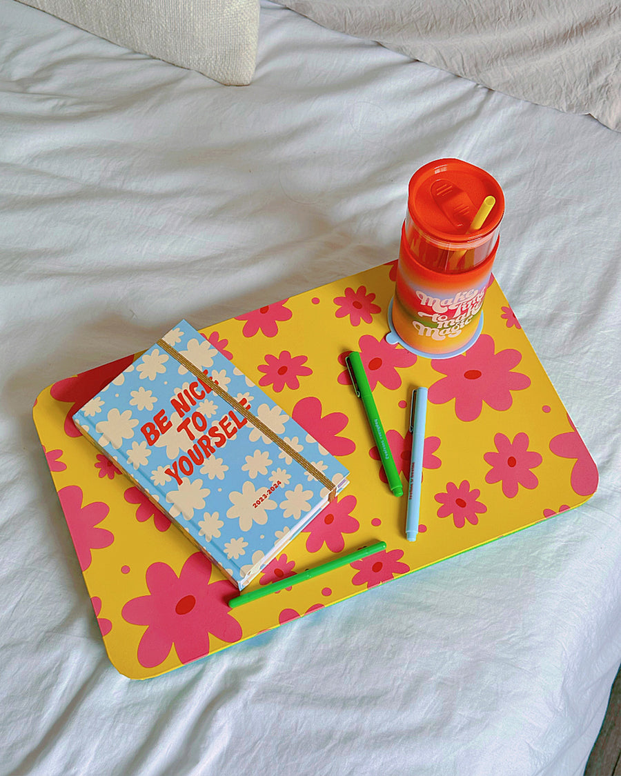 Yellow lap desk with pink flowers, sitting on a white bed with a planner.