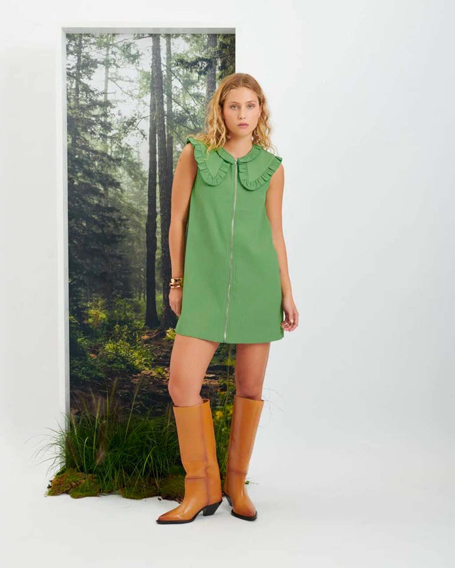 Woman in knee high brown boots standing in front of a woodland scene, wearing a green mini dress