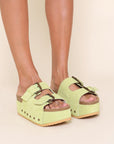 model wearing platform suede slide sandals with thick straps and metal buckle closures in pistachio green. shown from shin down