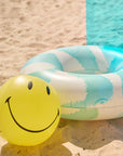 blue and white tye dye pool ring with smiley face graphic with matching yellow smiley face beach ball on a beach