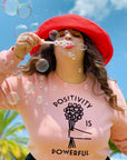 woman blowing bubbles wearing a pink sweatshirt with "Positivity is Powerful" text graphic and red hat