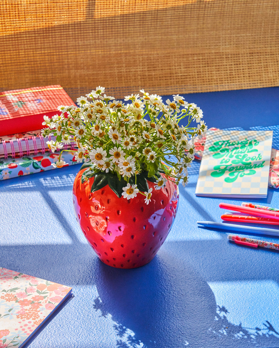 strawberry vase with white flowers on blue surface with desk accessorie scattered throughout