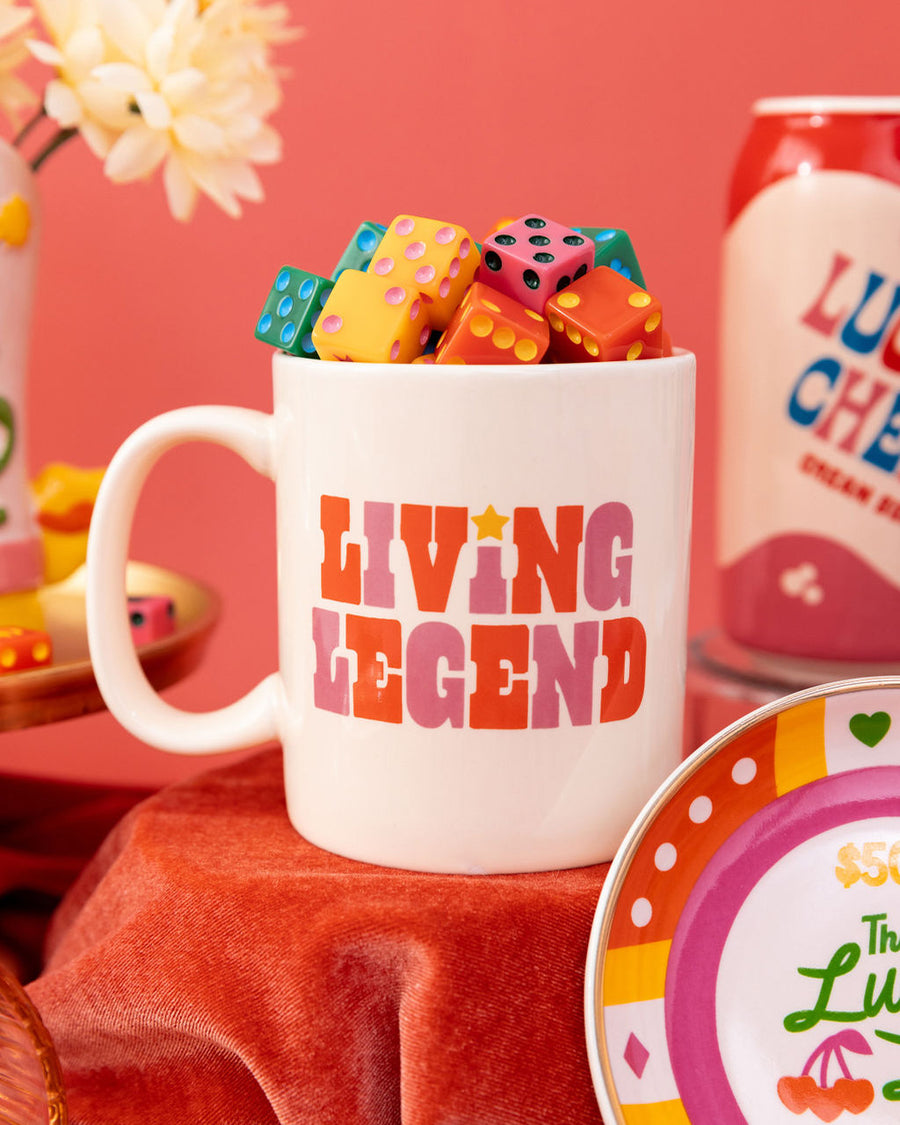 editorial image of ceramic mug with white ground and link and red 'living legend' across the front filled with colorful dice