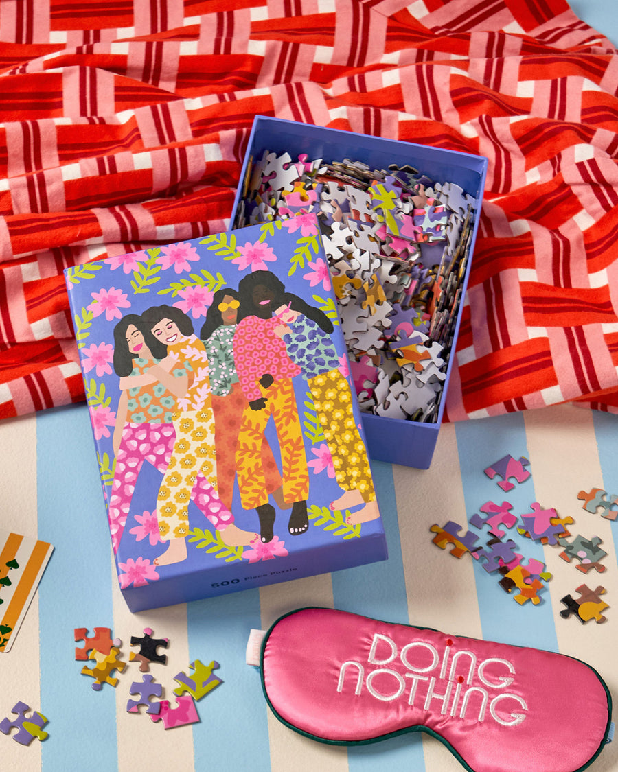 open puzzle box of illustration of 5 girls in bold floral print outfits against floral pattern and a sleepmask with "DOING NOTHING" text graphic, shown against a blue and white striped backdrop with red basket weave pattern fabric